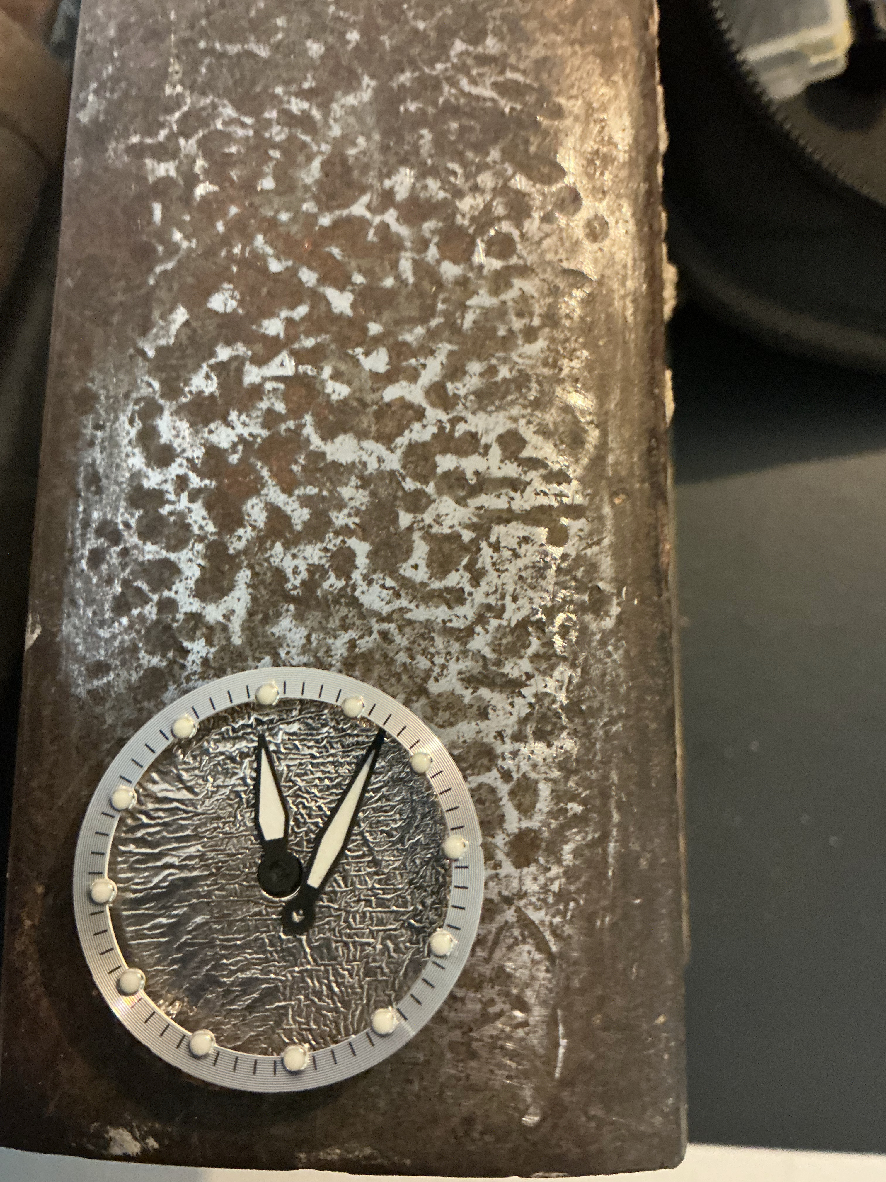 A reticulated silver dial with hands fitted, resting on a patterned steel block